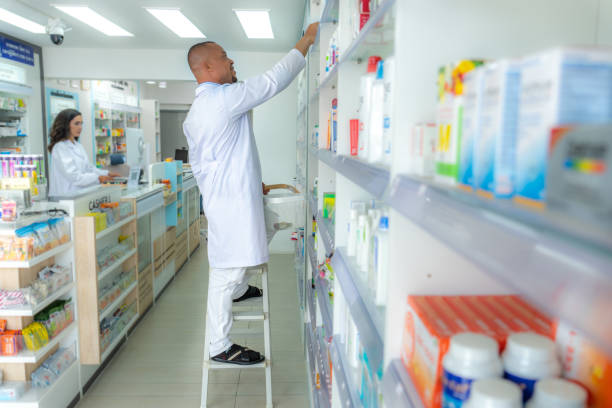 farmacéutico afroamericano pisando la escalera frente a los estantes de medicamentos para revisar las existencias de medicamentos mientras su colega latina está parada en el mostrador para preparar para dispensar medicamentos al cliente en la farmacia. - pharmacy pharmacist customer latin american and hispanic ethnicity fotografías e imágenes de stock