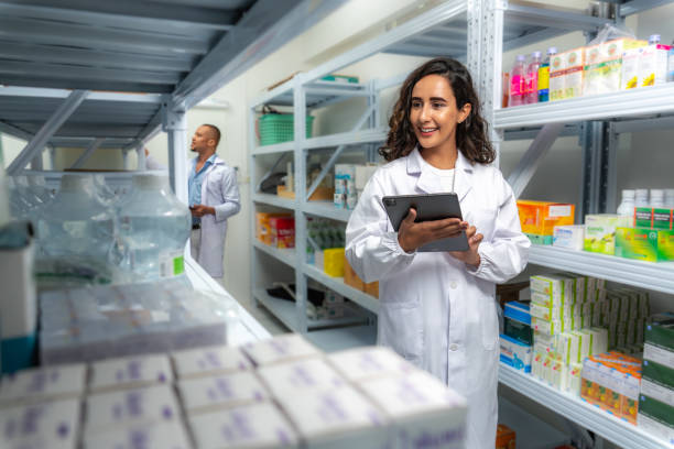 Beautiful Latin female startup entrepreneur, small business owner wearing white lab coat, holding digital tablet and checking medicine inventory together with her African man coworker at inventory room. hermosa empresaria latina de inicio, propietaria de una pequeña empresa con bata blanca de laboratorio, sosteniendo una tableta digital y revisando el inventario de medicamentos junto con su compañero de trabajo africano en la sala de inventario.
