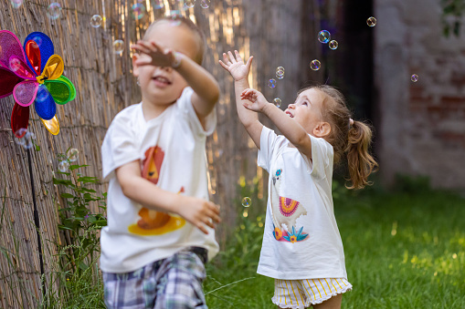A young boy and girl giggling as they blow bubbles together in the yard.