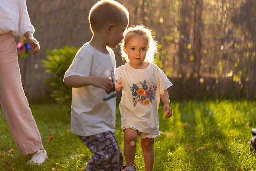 Two adorable siblings, a brother and sister, running hand in hand through a vibrant green backyard.