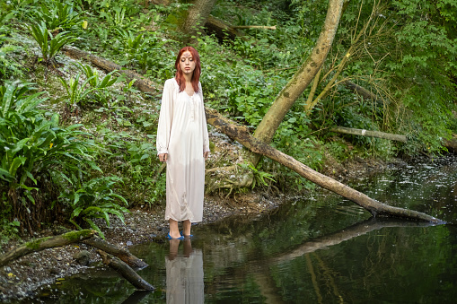 A young teenage girl in a white dress stands in a lake against a background of green foliage. day Ivan bathed.