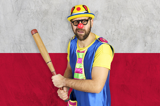 An insecure clown holds a bat in his hands against the background of the flag of Poland.