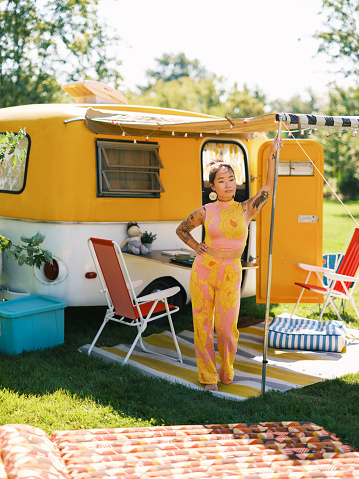 Mid adult Asian woman enjoying trailer camping in summer. She is dressed in retro casual outfit. Exterior of camping trailer in summer.