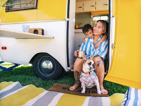Asian mother, son and small dog enjoying camping at the trailer site. Both dressed in casual retro clothing. Camping trailer is vintage style from 1974. Exterior of camping site on a sunny day.