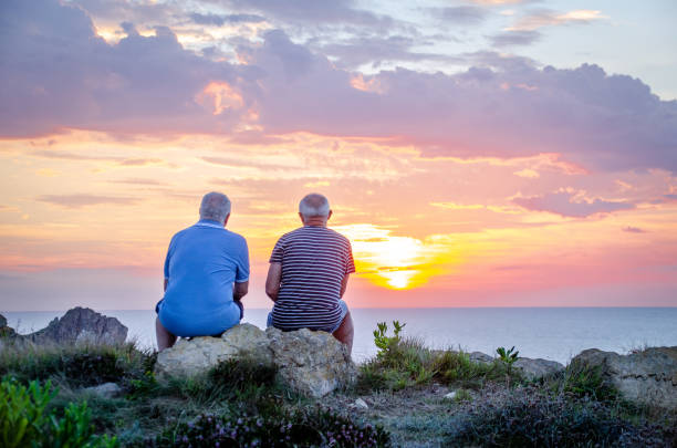 zwei alte männerfreunde von hinten gesehen und betrachten den schönen sonnenuntergang, der in den felsen einer bergklippe vor dem meer sitzt - contemplation sunset eternity thinking stock-fotos und bilder