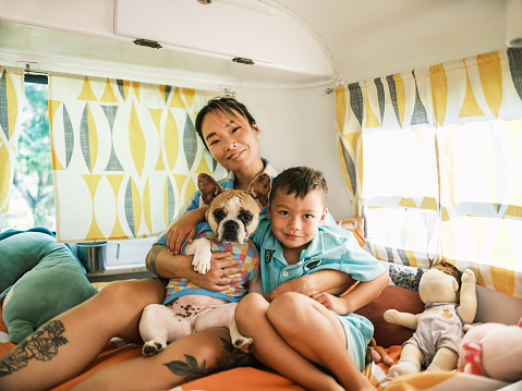 Asian mother, son and small dog playing on bed inside camping trailer. Both dressed in casual retro pyjamas. Camping trailer is vintage style from 1974. Interior of vintage trailer on a sunny day.
