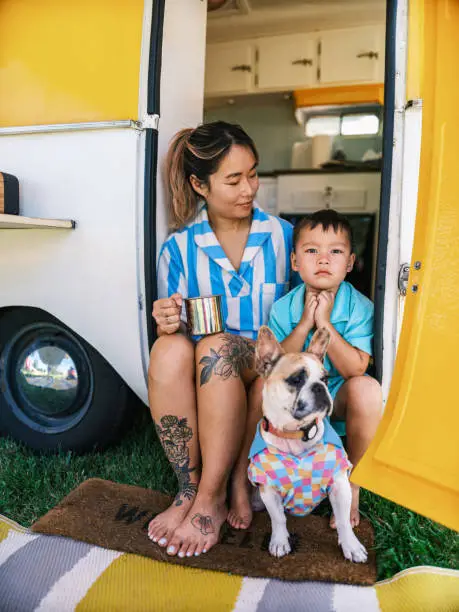 Asian mother and son enjoying camping with small French Bulldog at the trailer site. Both dressed in casual retro clothing. Camping trailer is vintage style from 1974. Exterior of camping site on a sunny day.