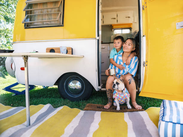 Asian mother and son enjoying camping with small French Bulldog at the trailer site. Both dressed in casual retro clothing. Camping trailer is vintage style from 1974. Exterior of camping site on a sunny day.
