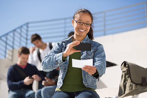 Young woman uses smartphone to deposit a check Happy young female student depositing check through mobile phone bank deposit slip stock pictures, royalty-free photos & images