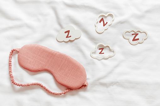 Above view pink sleep mask and clouds shapes with zzz snore sounds on white wavy cloth background. Eye cover mask for best sleep. Concept dream well, comfort rest at night. Minimal Aesthetic style