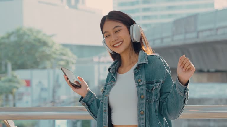 Young asian woman using headphone for listening nice music and standing in city. Girl happiness, smile face. Female love listen music that make her relaxed and enjoying life