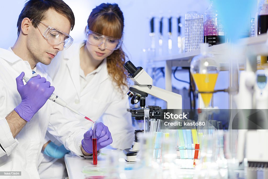 Health care professionals testing chemicals in a lab Young male researcher pipetting red liquid in the glass tube in the (forensics, microbiology, biochemistry, genetics, oncology...)laboratory. Female asistant science student watching and learning the protocol. Adult Stock Photo