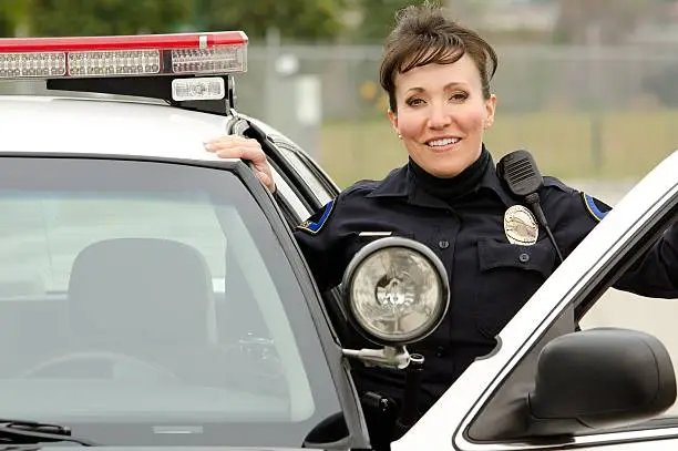A Hispanic female police officer smiles while standing next to her patrol car.