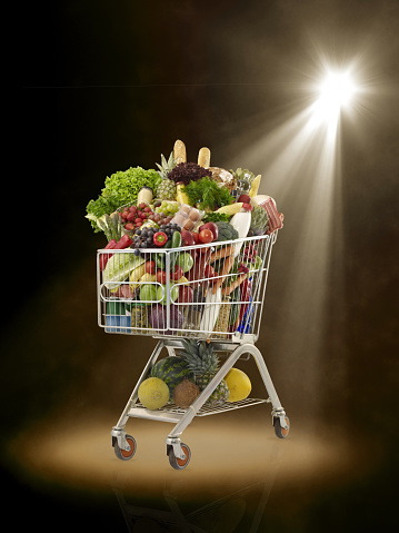 Shopping cart full of fresh vegetables, fruits and groceries under a spotlight ın a colored background stage