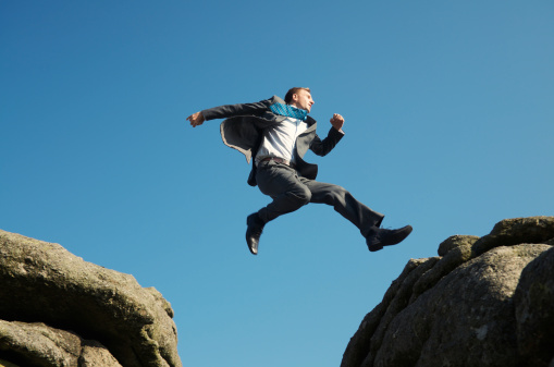Energetic young man businessman jumping outdoors between rocks high in bright blue sky