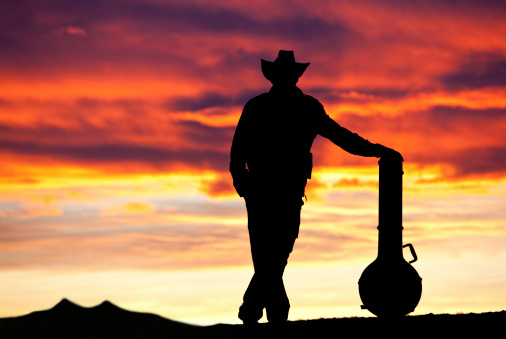 A silhouette of a country musician standing with a banjo. Cowboy. Dramatic sunset. Relaxed. Unrecognizable male roots music singer songwriter with banjo and acoustic instrument standing against an amazing sunset. Wearing cowboy hat. Themes include country music, roots music, poet, guitar player, guitar, banjo, stringed instrument, bluegrass, country music, outside, festival, music festival, arts entertainment, and guitars. Male singer is unrecognizable. 