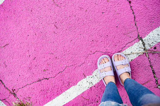 This is a photograph shot from a female perspective looking down past jeans with feet wearing purple sandals while standing on a painted line.