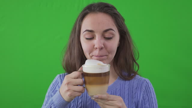 Close-up clicking clapperboard and Caucasian beautiful woman drinking coffee looking at camera. Front view portrait of confident charming actress advertising cappuccino at green screen background.