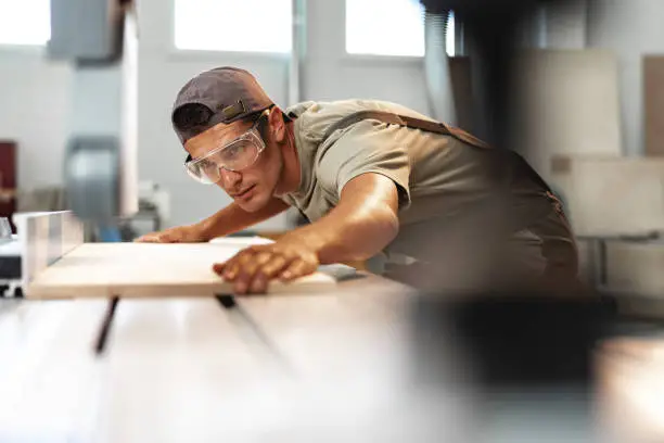 Photo of Young carpenter cutting a piece of wood in using a circular saw in furniture factory