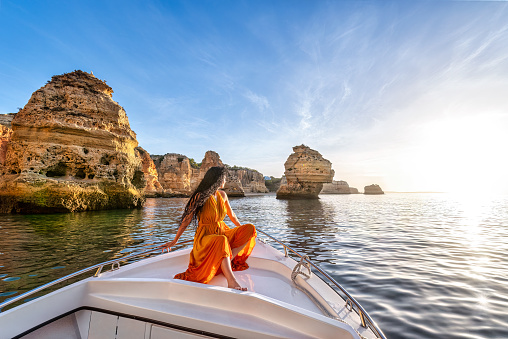 Portuguese woman with long hair and orange dress sailing on the yacht near the rocky coast of Algarve near Benagil Cave, Praia da Marinha and Carvoeiro in the morning, Portugal