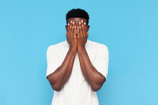 african american man in white t-shirt is shy and afraid on blue isolated background, guy in stress covers his face with his hands and hides