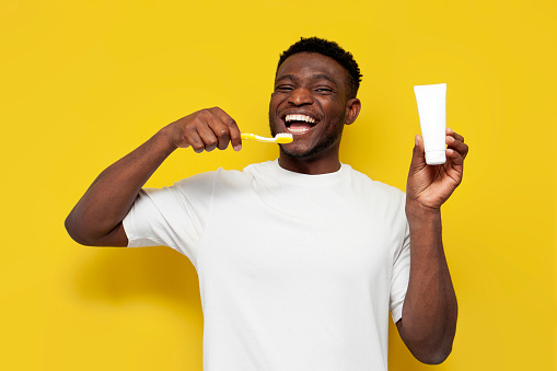 joyful african american man brushing his teeth and holding toothbrush and tube of toothpaste on yellow isolated background, the guy takes care of his teeth and smile