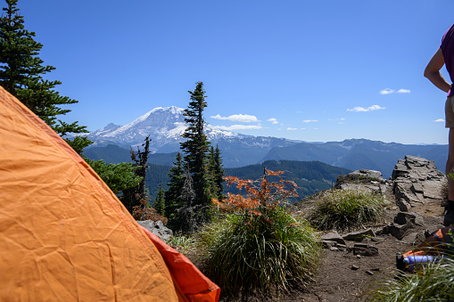 An orange tent and a young woman by the Summit Lake trail with Mount Rainier in the distance. Camping at Mt Rainier National Park. Washington State.