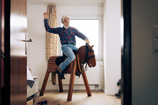 Indoor portrait of senior man playing cowboy in grandchild's room, sitting on wooden horse.