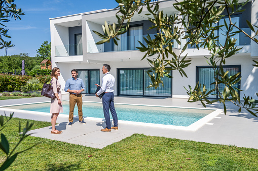 Male real estate agent talking with couple while standing in front of modern house during sunny day.
