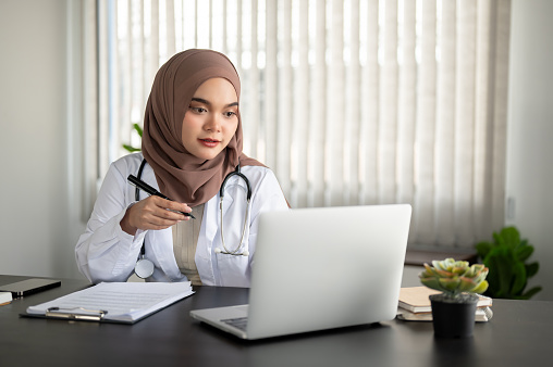 A beautiful and professional Asian Muslim female doctor in a uniform and hijab is working on her laptop at her desk in the office at a hospital or clinic.