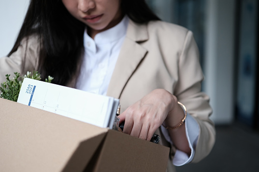 Frustrated fired female employee packing belongings in cardboard box leaving workplace. Unemployment concept.