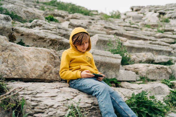 dziecko w wieku szkolnym bawi się smartfonem z telefonu komórkowego w starożytnym kamiennym amfiteatrze. chłopiec w żółtej bluzie z kapturem wysyłający sms-y przez gadżet telefonu, siedząc na kamiennej ławce w opuszczonej zabytkowej arenie - ancient civilization people time visual art zdjęcia i obrazy z banku zdjęć