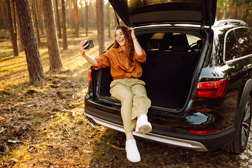 Happy stylish female tourist in the trunk of a car with a phone, taking a selfie, talking on a video call. A woman in the trunk of a car at sunset. Traveling by car, camping. Active lifestyle.
