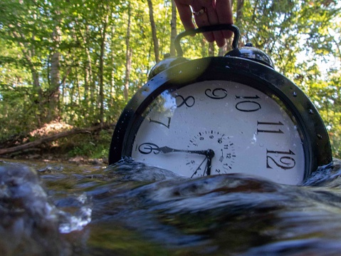 Concept image losing track of time, time running out relativity science image, flowing time, large clock in a flowing stream with half of the clock face submerged with ripples and colors distorting the numbers. Ouitdoors with no people and copy space.