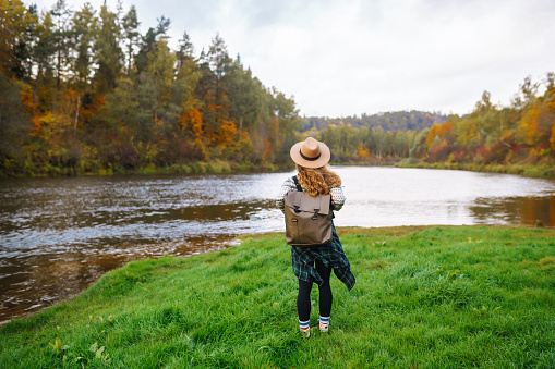 A tourist in a stylish sweater and hat with a backpack admires the view of the river. A young girl in a hat enjoys nature. Travel concept. Autumn mood.