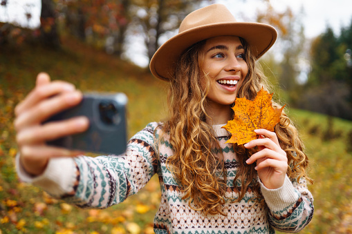 Beautiful woman in a stylish sweater with a phone in her hands sits in a clearing among yellow fallen leaves in an autumn park. Tourist enjoys the weather, takes a photo with a yellow leaf.