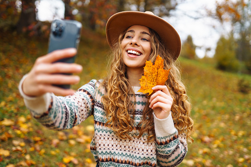 Beautiful woman in a stylish sweater with a phone in her hands sits in a clearing among yellow fallen leaves in an autumn park. Tourist enjoys the weather, takes a photo with a yellow leaf.