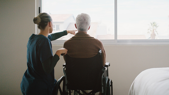 Senior man, wheelchair and caregiver pointing at view in bedroom for relax, conversation and care in nursing home. Wellness, professional woman and elderly person at window for nature and lifestyle