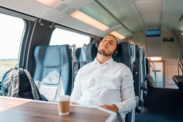 A young Caucasian man is sleeping on a train with his headphones on his head.