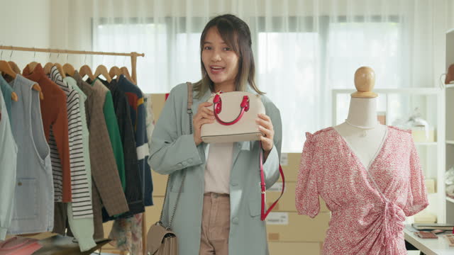 Young Asian woman live streaming on phone show new collection handbag to customer at shop. SME small business e-commerce.