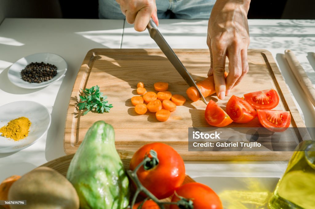 High angle of crop anonymous woman cutting carrot on wooden chopping board near tomatoes and parsley in kitchen during sunny weekend Vegetable Stock Photo High angle of crop anonymous woman cutting carrot on wooden chopping board near tomatoes and parsley in kitchen during sunny weekend Vegetable Stock Photo