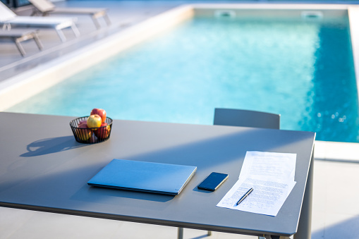 Laptop, mobile phones with documents on table at poolside during sunny day.