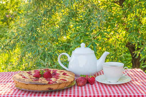 Cake with strawberries, white cup of tea and tea pot on the table covered by red and white checkered tablecloth