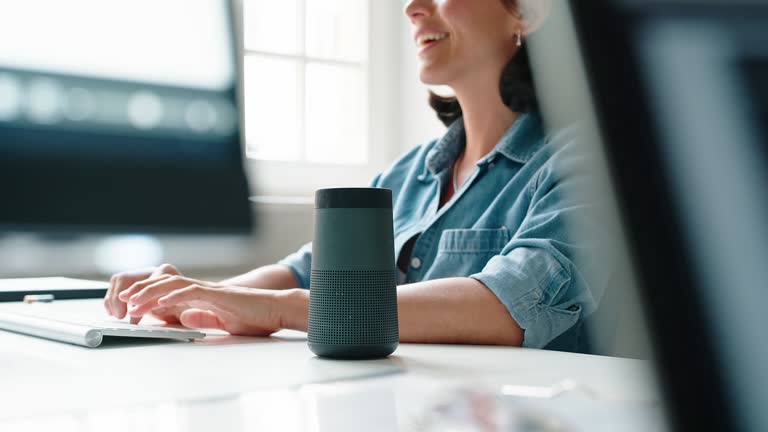 Woman business professional working on computer and talking to a virtual assistant speaker at startup office