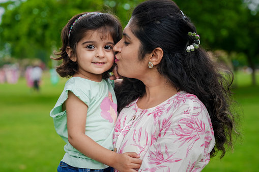 Happy young indian mother kiss her cute little girl daughter at summer park.