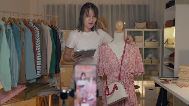 Young Asian woman live streaming on phone show new collection handbag to customer in shop at night. SME small business e-commerce.