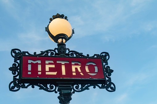 A metro sign for the Paris underground, with a light bulb on the top. Dusk. Blue sky in the background.