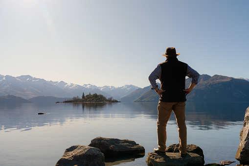 Sporting an Indiana Jones-style hat, a man in his fifties takes in the wonders of the still glacial waters of Lake Wanaka and Ruby Island. Beyond the island we see the towering snow-capped peaks of the Southern Alps.