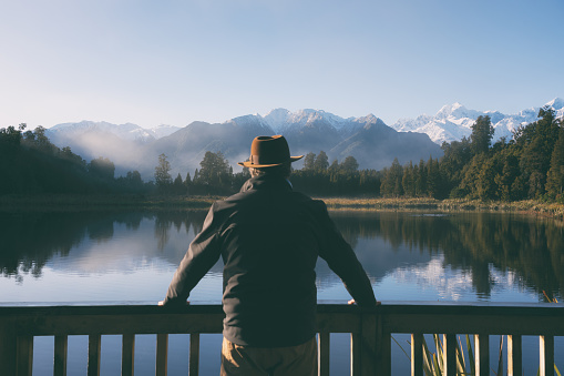 Sporting an Indiana Jones-style hat, a man in his fifties takes in the wonders of spectacular Lake Matheson on New Zealand’s South Island, and beyond we see the snow capped peaks of Mt Tasman,  Mt Cook and the Southern Alps.