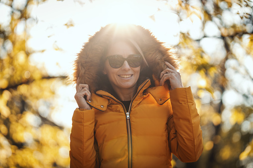 Beautiful young woman is holding jacket hood around her face in the autumn park and smiling.